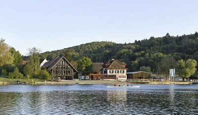 Hotel and Restaurant 1802+ located on the River Rhine, showing historic riverside buildings with outdoor terrace seating and small boats along the shore.