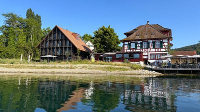 Hotel and Restaurant 1802+ located on the River Rhine, showing historic riverside buildings with outdoor terrace seating and small boats along the shore.