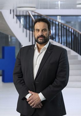 dark-haired man with beard in black suit, standing in a hall in front of blue GF cross and white stairs