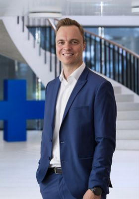 man in dark blue suit, standing in a hall in front of blue GF cross and white stairs