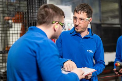 A vocational trainer is explaining something to an apprentice in the production hall.