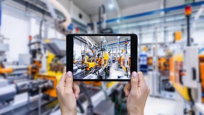 Horizontal color image of female hands holding a digital tablet in a modern plastic production line. Ordering on-line from injection moulding factory on a touchscreen tablet computer. Large factory, industrial machines, robots and manufacturing equipment arranged in background.