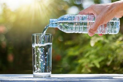 Hand holding drinking water bottle pouring water into glass on wooden table on blurred green nature background